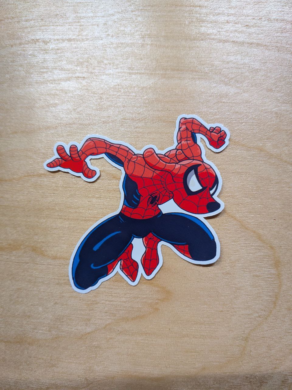A die-cut vinyl sticker of a stylized, cartoonish Spider-Man. The character is depicted with a rounder, stocky build in his classic red and blue suit with large white eyes. He is in a dynamic crouching pose with one hand on the ground and the on a light wood grain surface.