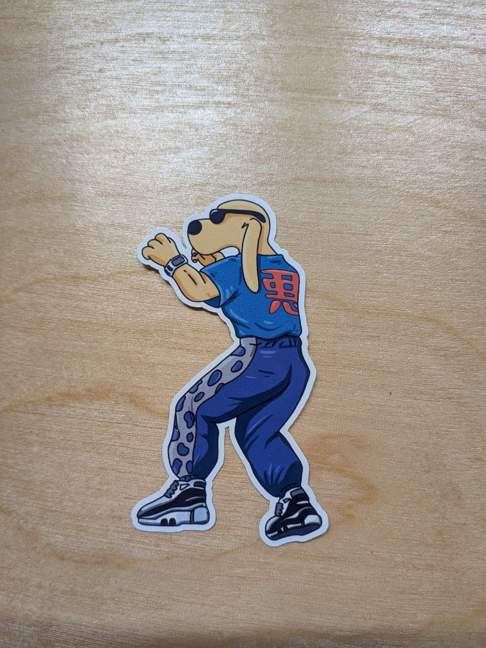 A die-cut vinyl sticker of a cartoon dog dressed as the Tekken character King in his casual outfit. The dog wears a light blue t-shirt with red Japanese text and purple track pants, resting on a wooden surface.